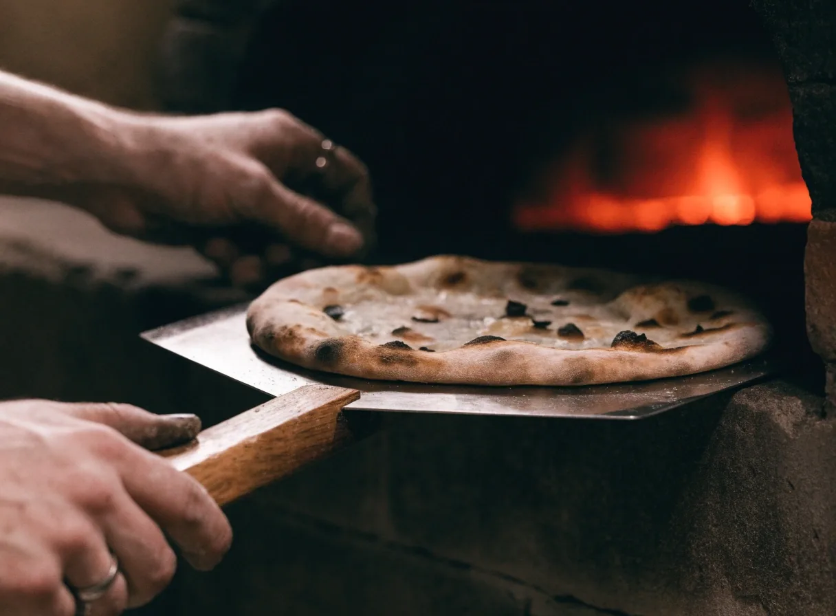 Hands guiding a pizza peel near a coal oven mouth